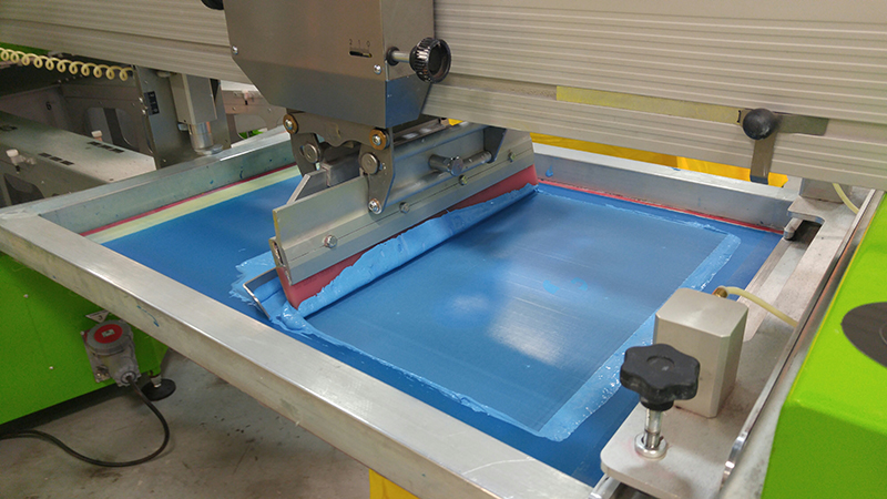 Screen-printing machine using blue ink to create a logo on customizable t-shirt.