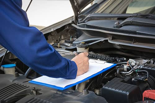 Mechanic writing on a notepad with the hood of the car open.